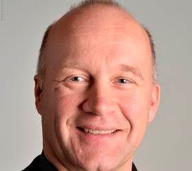 En skaldet mand med lys hud smiler bredt og vender fremad.