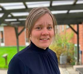 Kvinde med kort blond hår, smilende, iført mørkeblå rullekrave, udendørs.