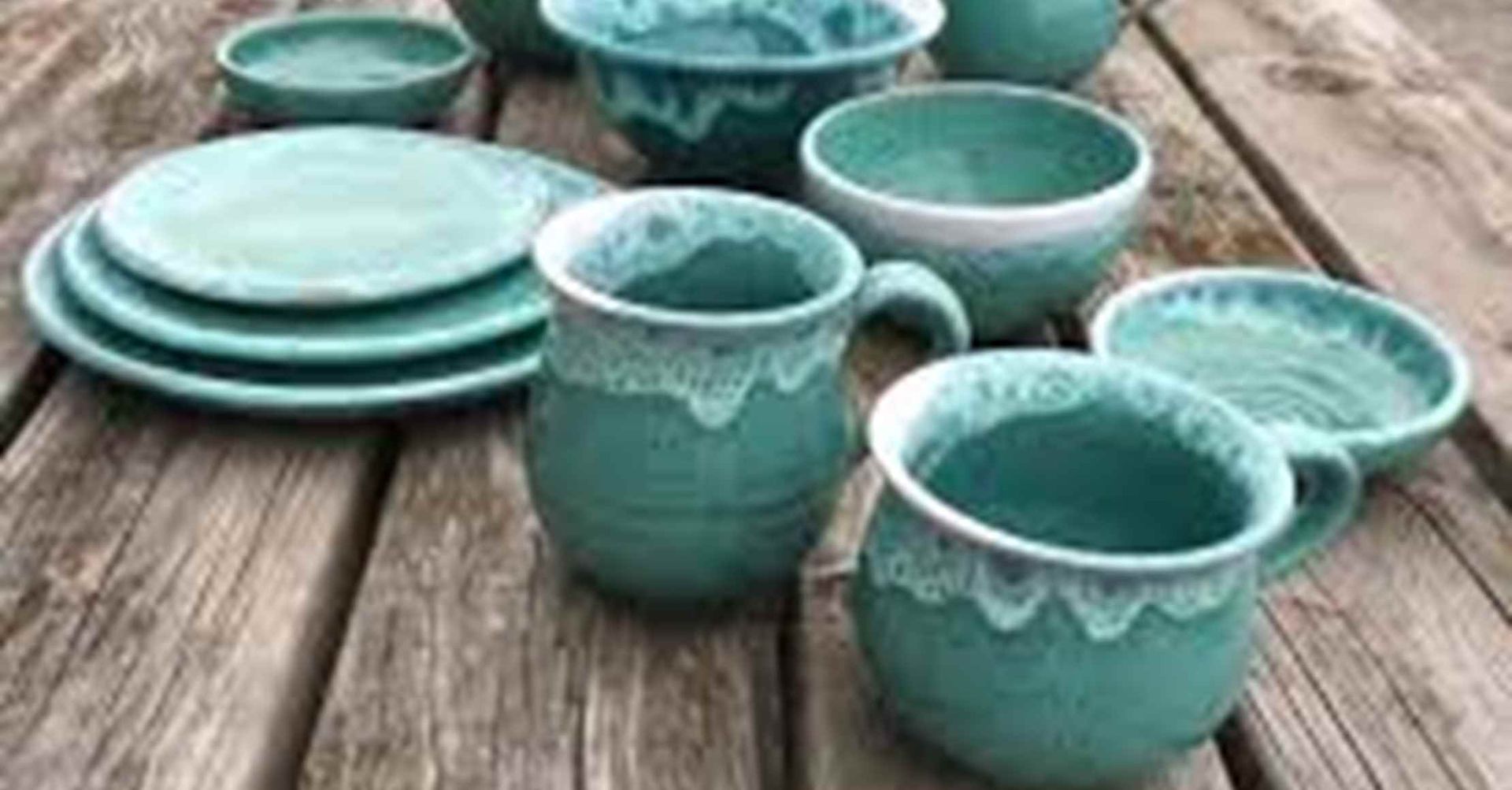 Keramisk service i blågrøn på en træoverflade; inkluderer tallerkener, skåle og krus med hvid glasurdetaljer.
