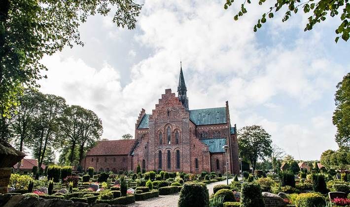Rød murstenskirke med et højt spir, indrammet af træer og en formel have, under en overskyet himmel.