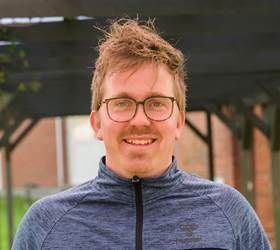 En mand med briller smiler, står udendørs foran en skyggefuld bygning, iført en blå skjorte med lynlås.