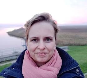 Kvinde med kort blond hår og lyserødt tørklæde smiler udendørs, vand og land i baggrunden.