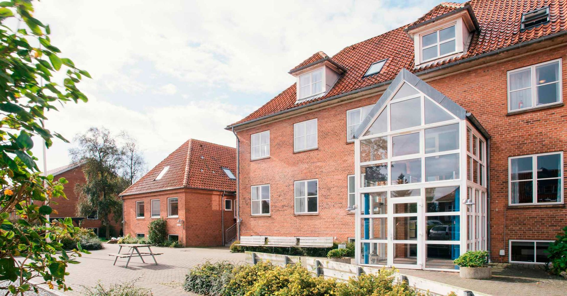 Rød murstenskirke med et højt spir, indrammet af træer og en formel have, under en overskyet himmel.