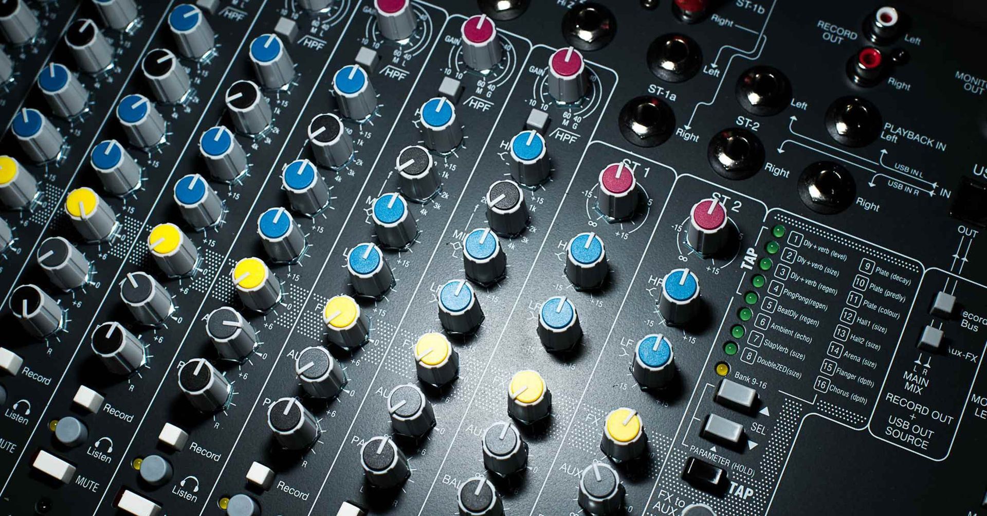 Nærbillede af en mikserpult med adskillige drejeknapper og knapper i forskellige farver på en mørk overflade.