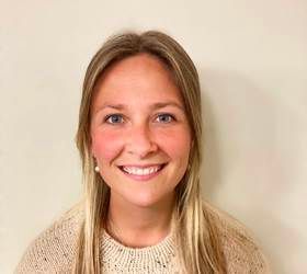 En blond kvinde smiler, iført en lys sweater, mod en ensfarvet baggrund.