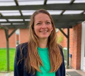 Kvinde med langt brunt hår smiler udendørs, iført en grøn skjorte og marineblå blazer.