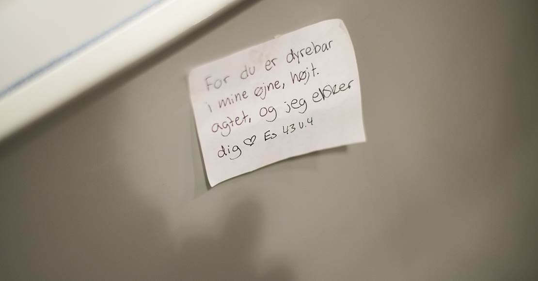 En håndskrevet seddel, tapet fast på en grå overflade, indeholdende tekst.