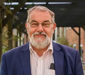 Mand med skæg og briller iført blå blazer, smilende udendørs.