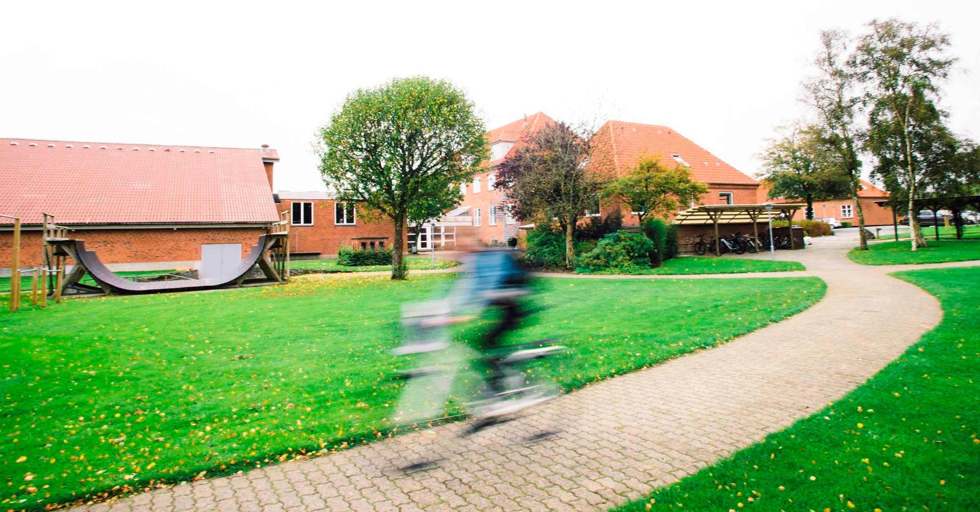 En cyklist sløret på en murstenssti gennem en grøn park, med bygninger med røde tage i baggrunden.