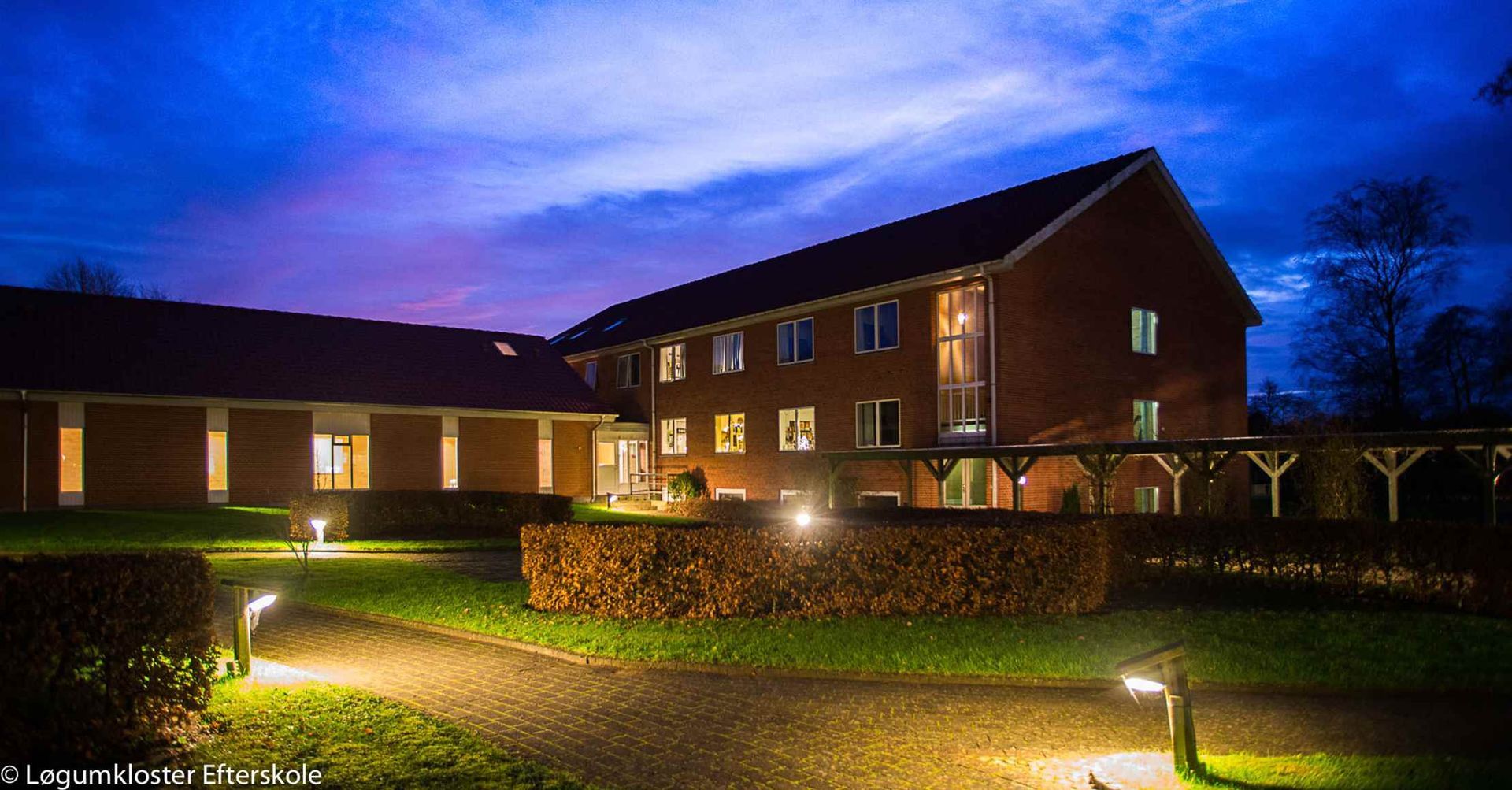 Murstensbygning i skumringen, oplyste stier, velplejet græsplæne og mørkeblå himmel.