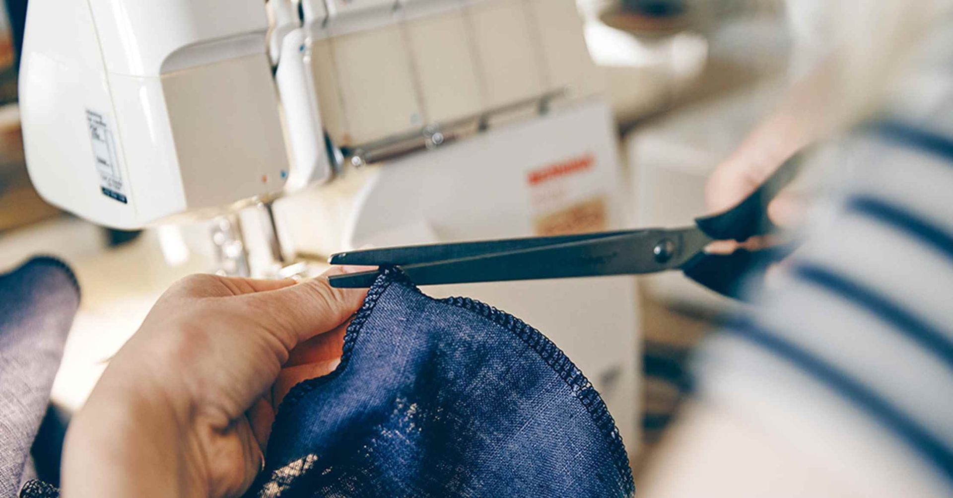 Person klipper stof med saks nær en symaskine. Blåt stof og hvid maskine er synlige.