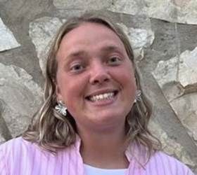 En kvinde med blond hår smiler, iført en lyserød skjorte, foran en stenmur.