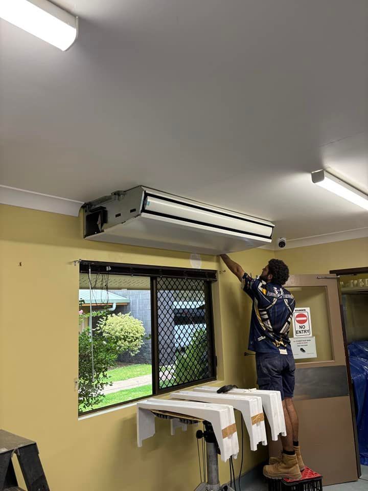 A person on a step stool installs an air conditioner on a yellow wall next to a window. — Active Airconz in Winnellie, NT