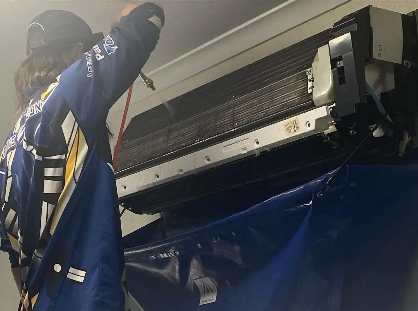 Person in Blue Jumpsuit Cleans an Air Conditioner With a Spray Nozzle, Indoors — Active Airconz in Winnellie, NT