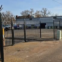 A metal fence surrounds a parking lot with a building in the background.