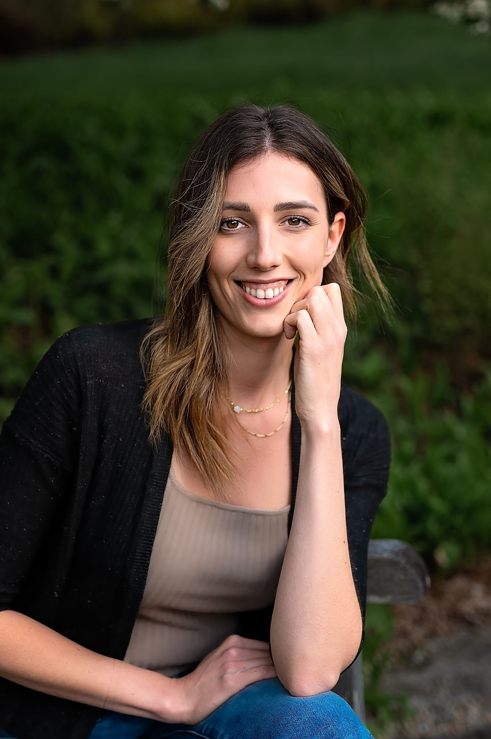 Woman smiles, resting chin on hand, wearing black cardigan over a tan top and jeans, outdoors.