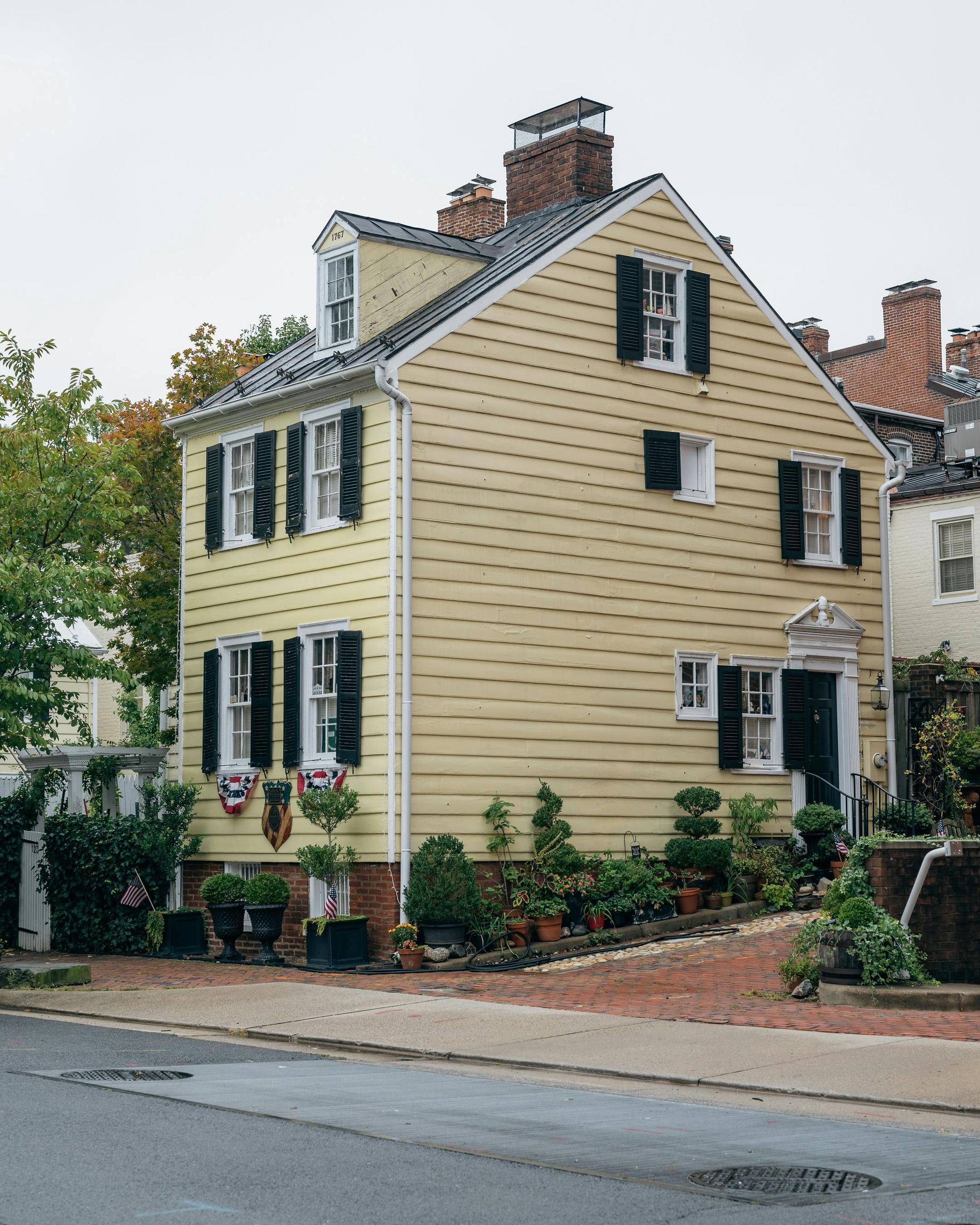 Yellow historic house with black shutters, potted plants, and brick path.