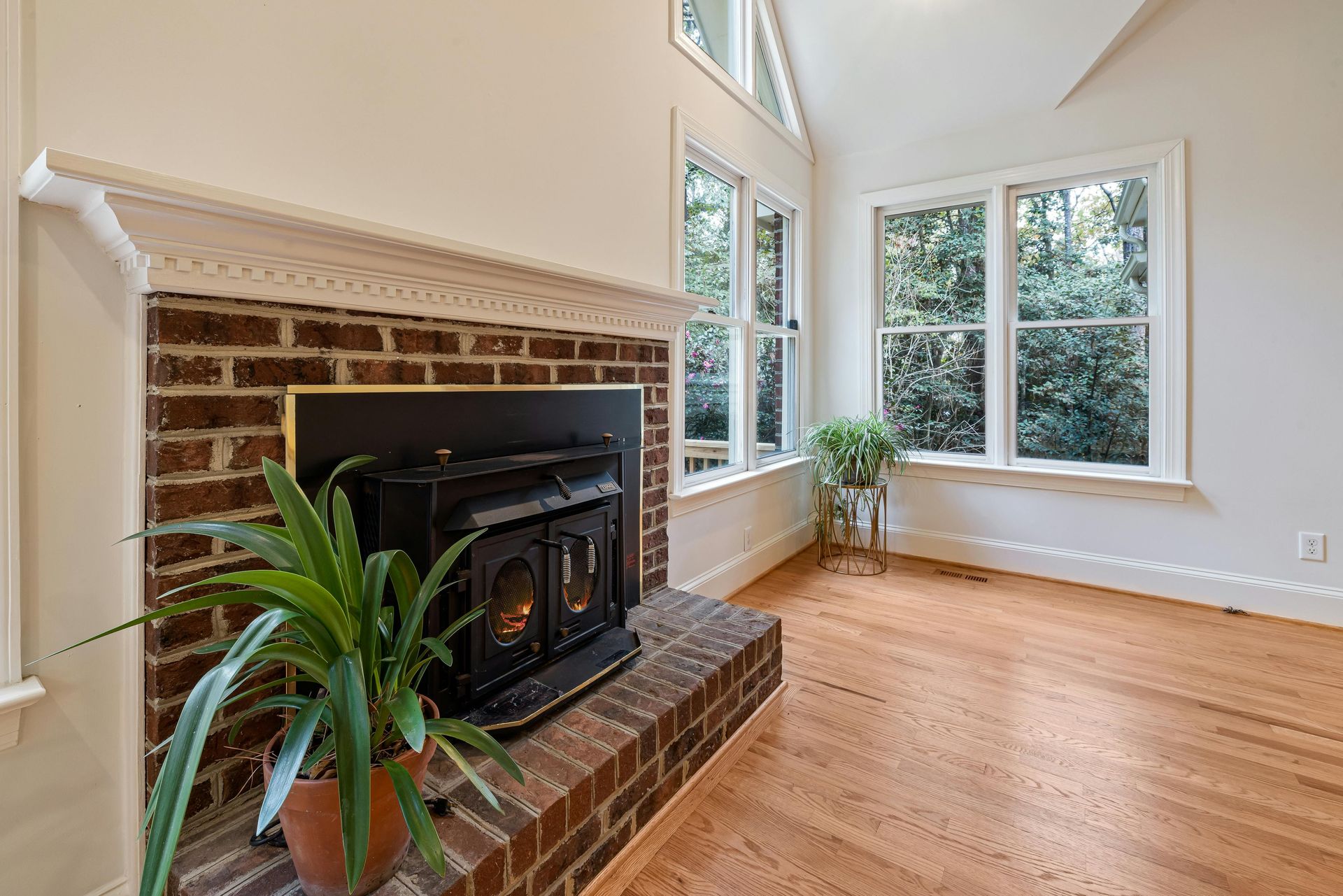 Living room with brick fireplace, wood floor, and large windows looking out to trees.