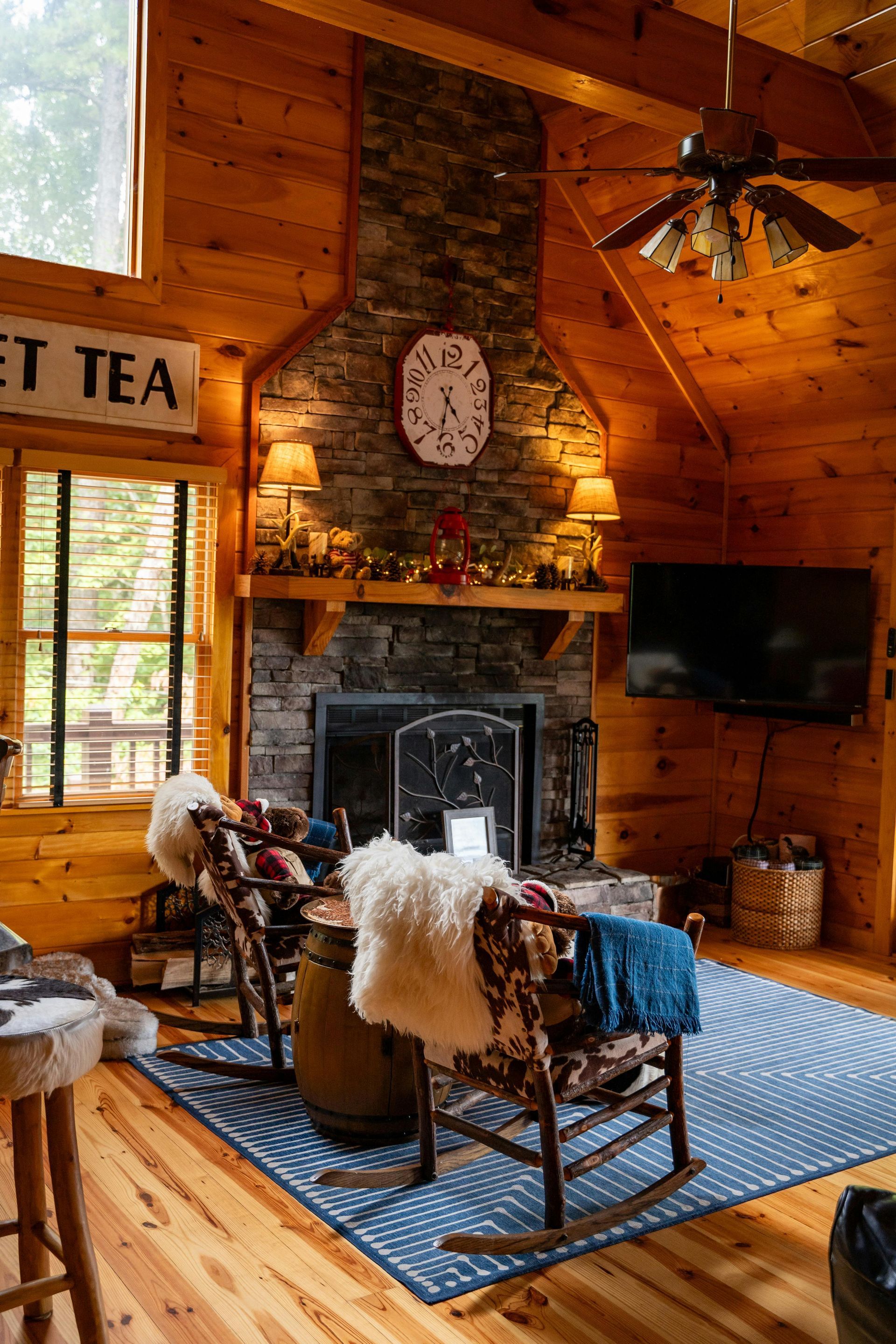 Cozy cabin interior with fireplace, rocking chairs, and TV. Wooden walls, blue rug, and clock above the mantle.