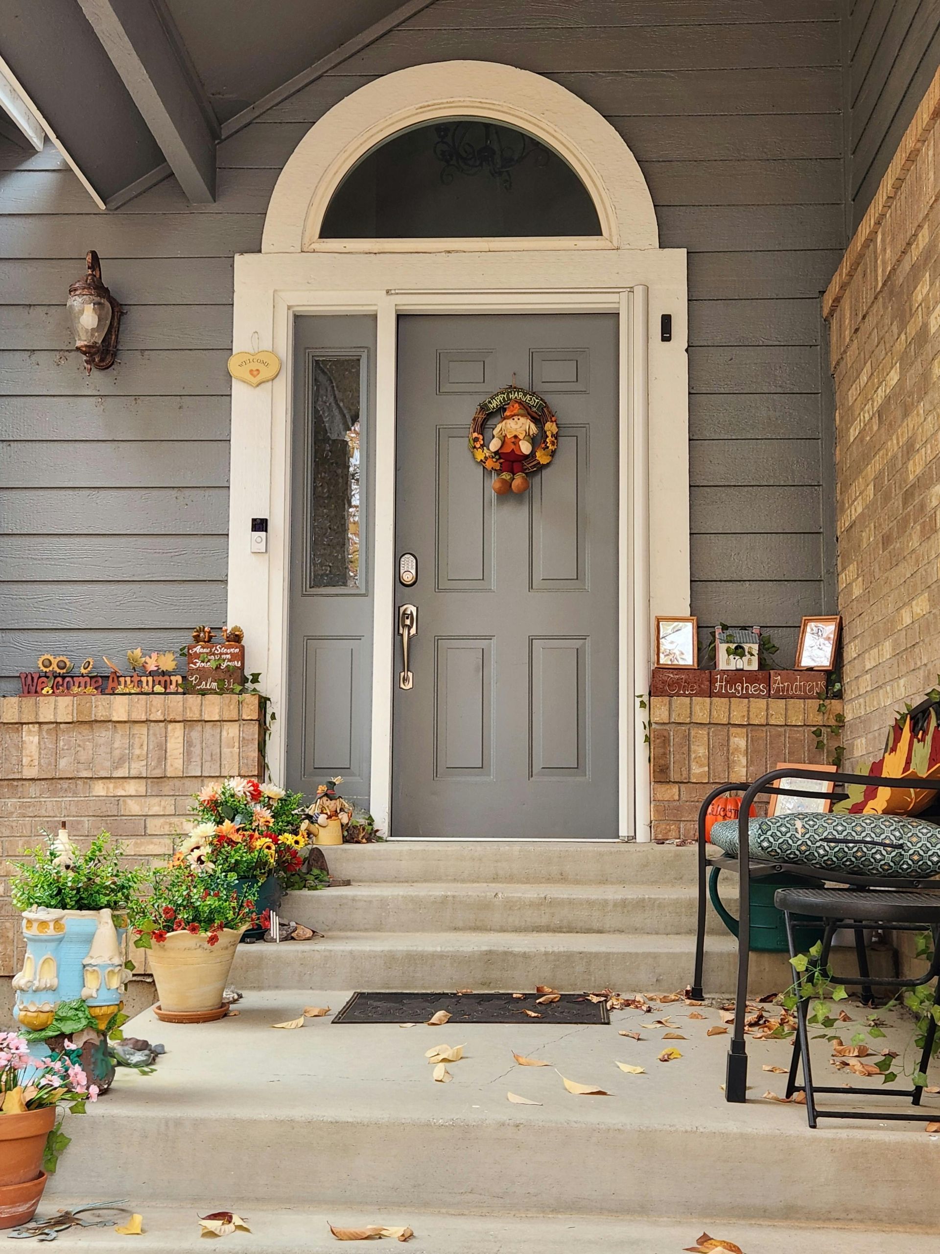 Gray front door with wreath and side window, on porch with flowers and steps.