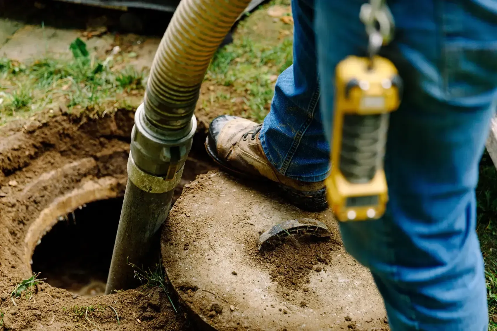 A person vacuums a septic tank through an open access. A yellow monitoring device hangs from their belt.