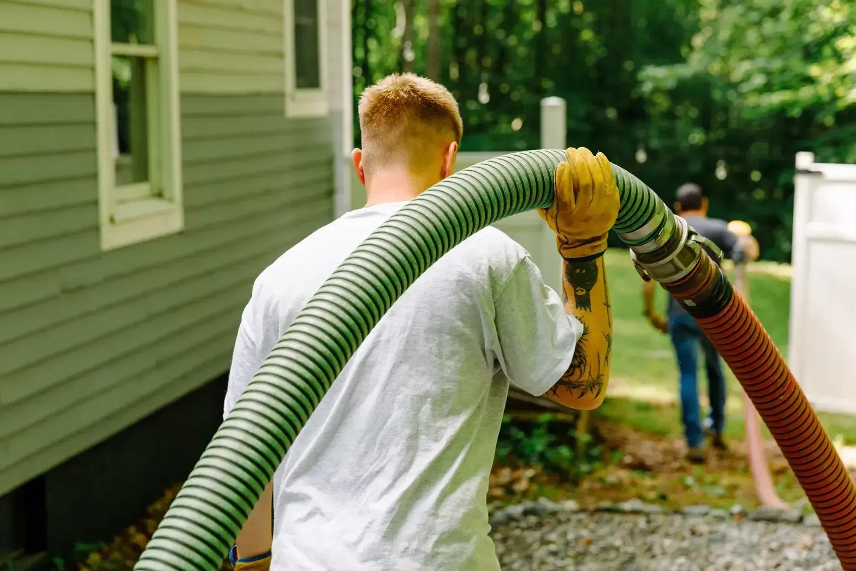 Man carrying a large hose near a house, another person walks behind him outdoors.