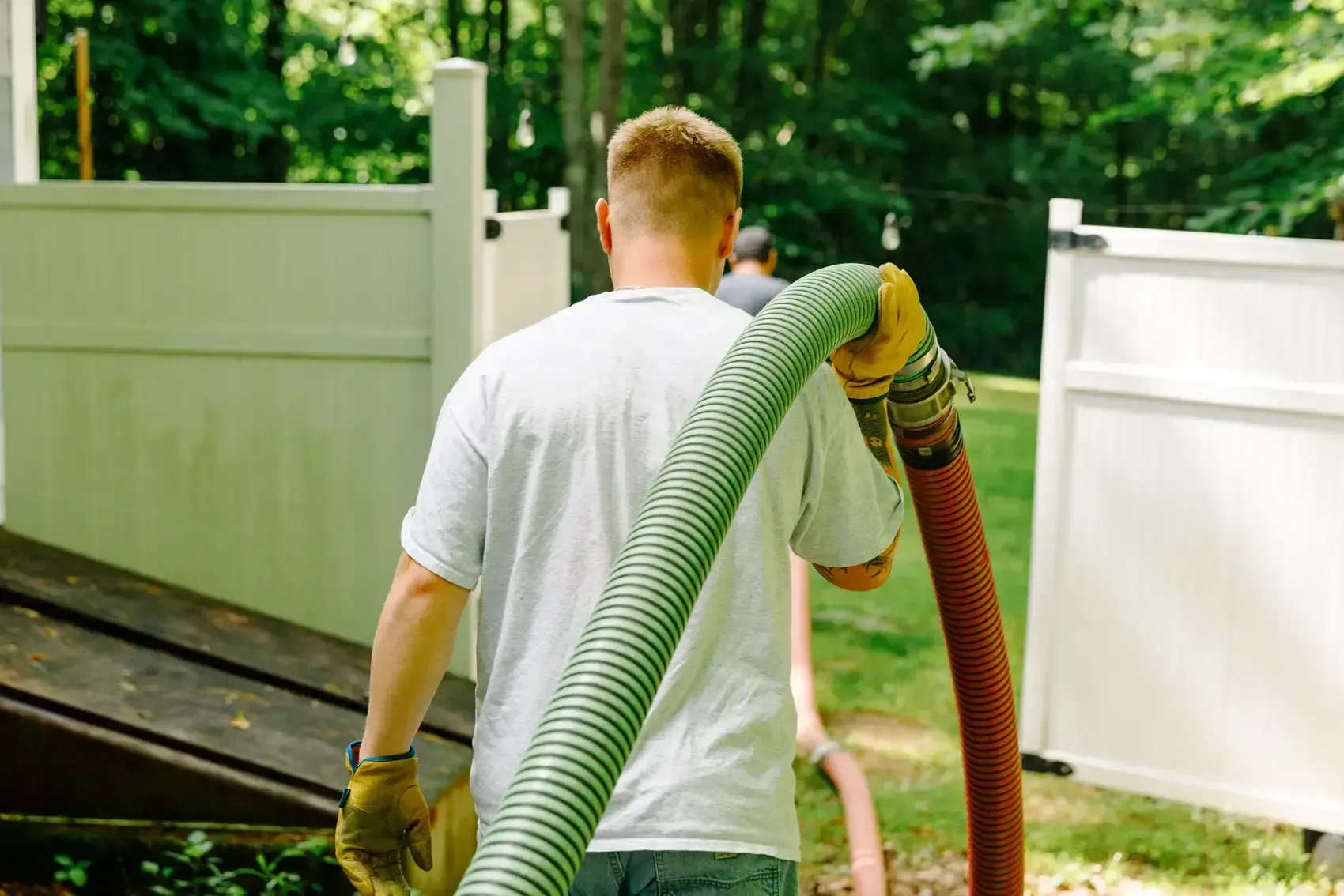 Man carrying a large hose through a fenced yard, another person walking ahead.