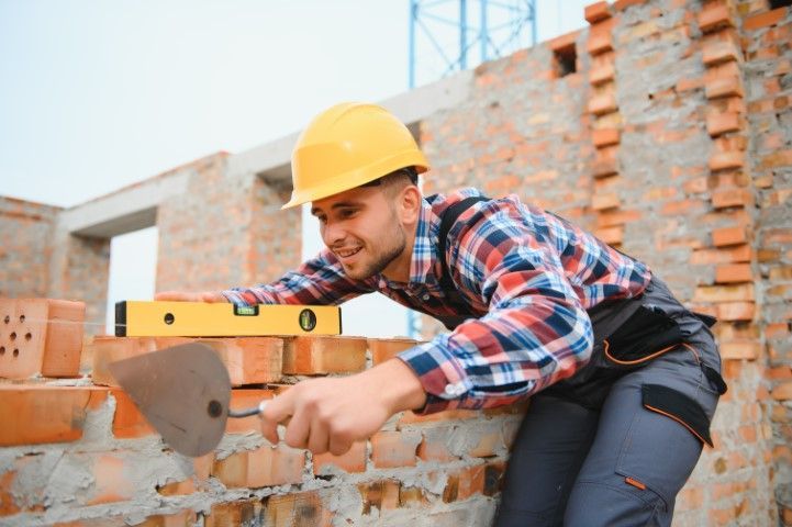 Construction worker in yellow hard hat using a trowel and level to lay bricks on a building site.