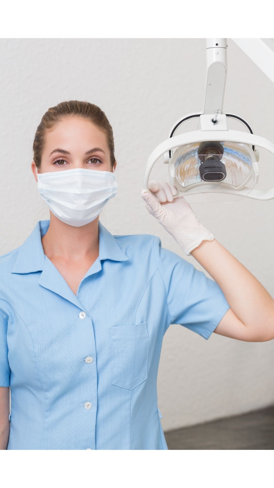A female dentist wearing a mask and gloves is holding a light.
