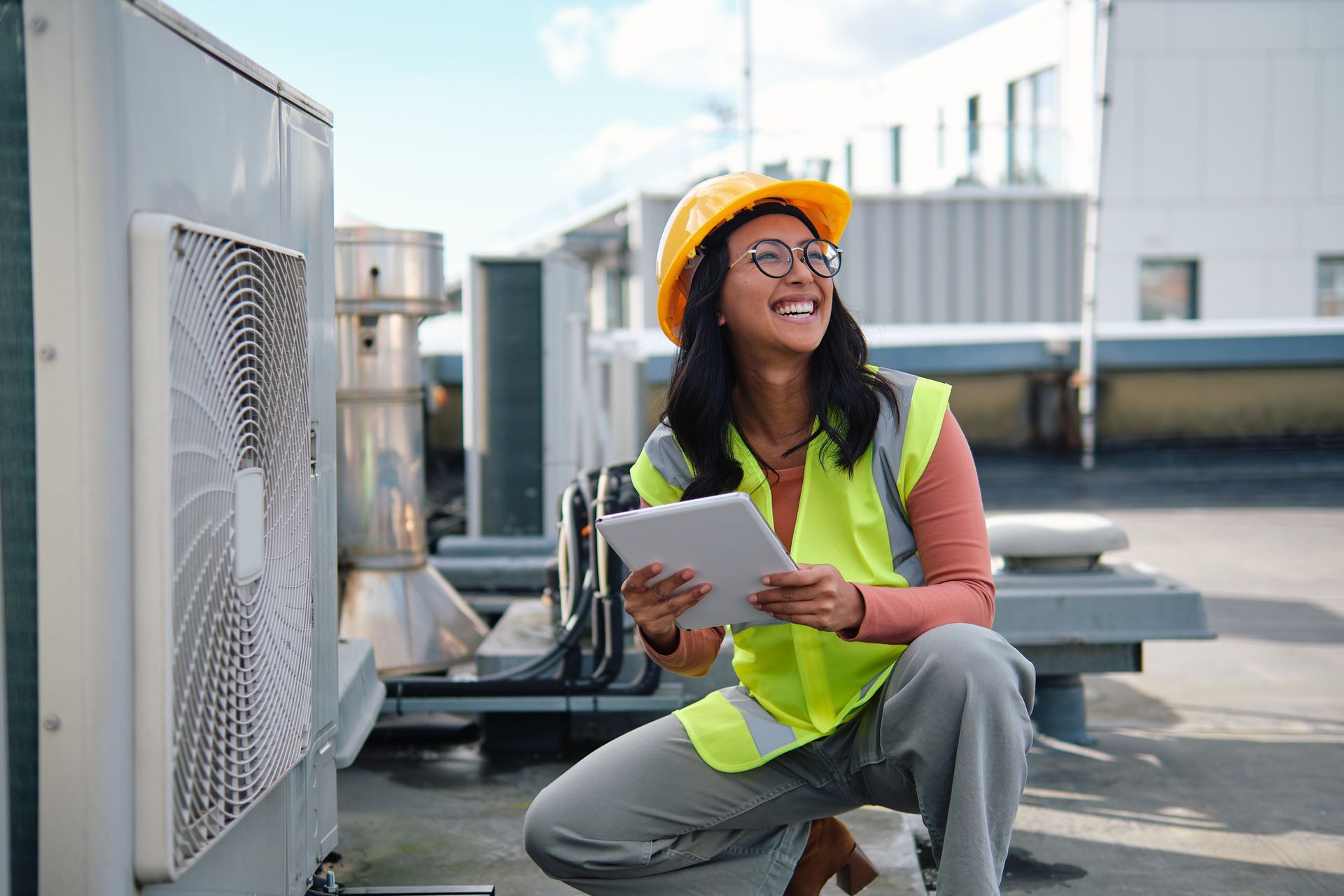 Woman in safety vest and hard hat inspects HVAC unit, using a tablet on a rooftop.