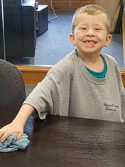 A young boy is smiling while cleaning a table with a cloth.