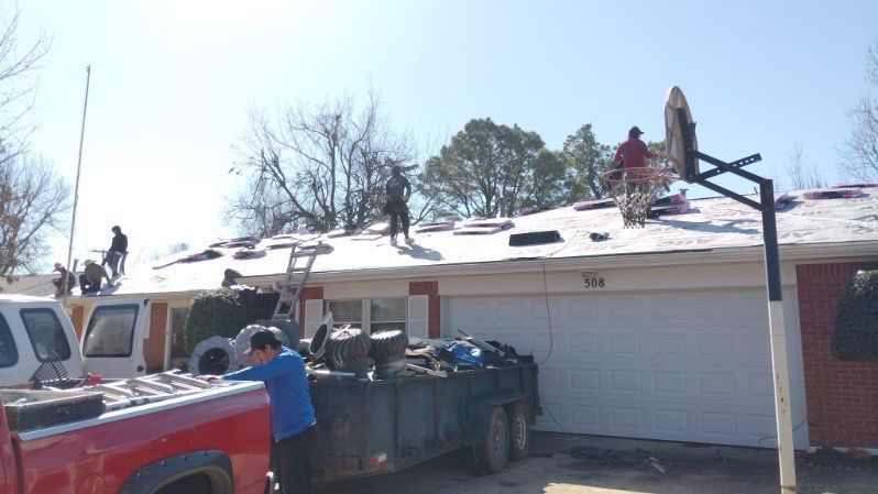 Roofers working on a house, removing old shingles. Crew members on roof, truck and trailer in yard.