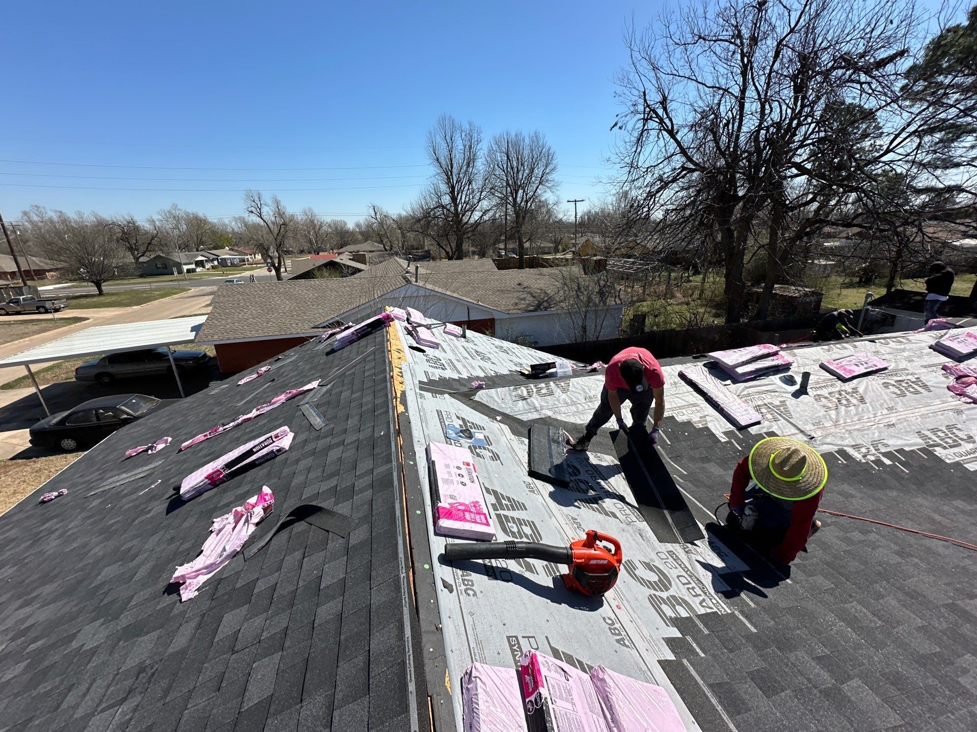 People roofing a house with new shingles; one uses a blower, and pink insulation is visible.