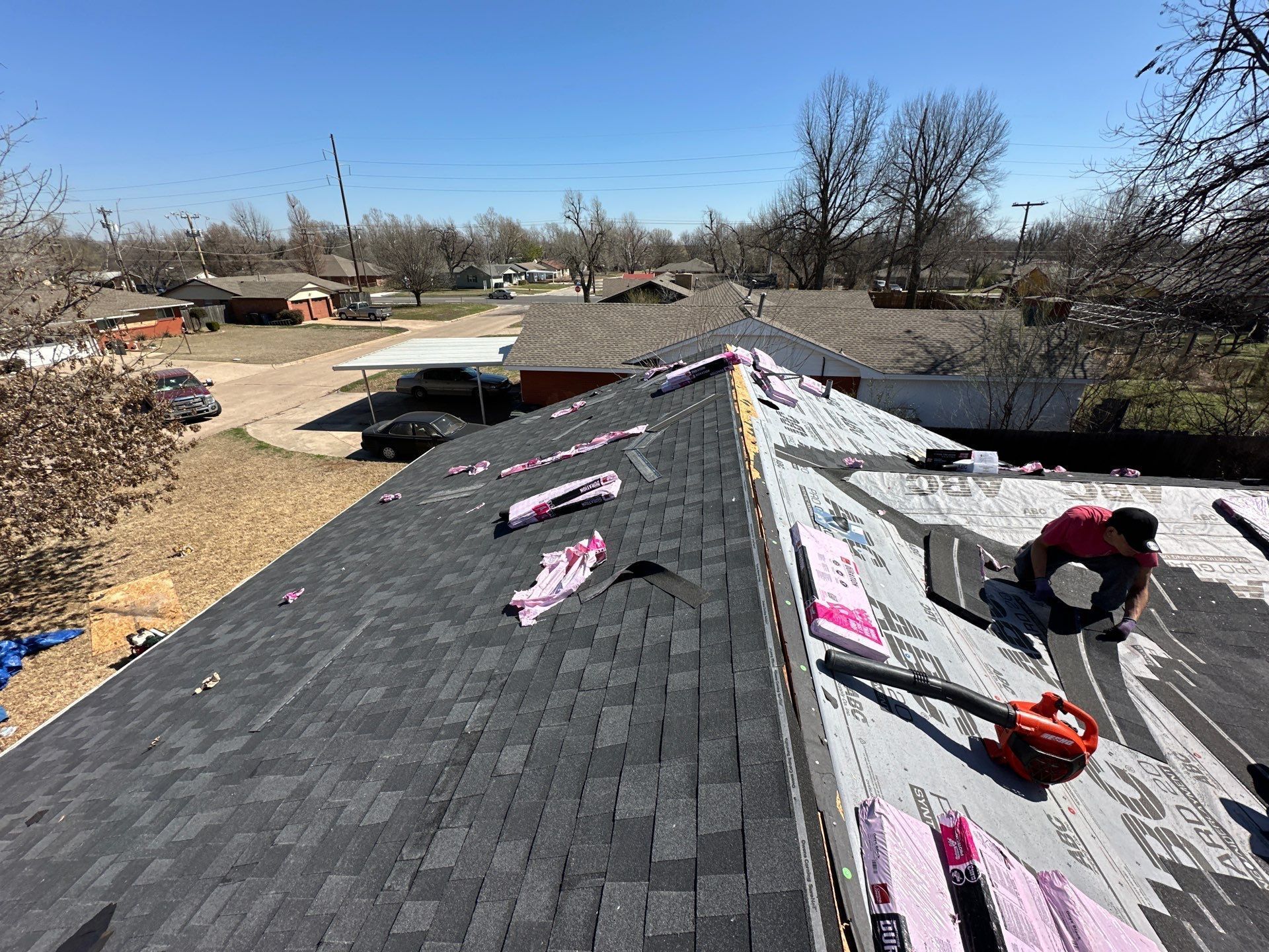 Roofer working on a roof, clearing debris with a blower. Buildings and a clear blue sky are in the background.