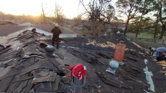 Workers repairing a damaged asphalt shingle roof, likely after a storm. Sun is setting, casting light on the scene.