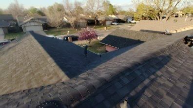 Overhead view of several dark-shingled roofs of houses, with a tree in the yard and other houses in the background.