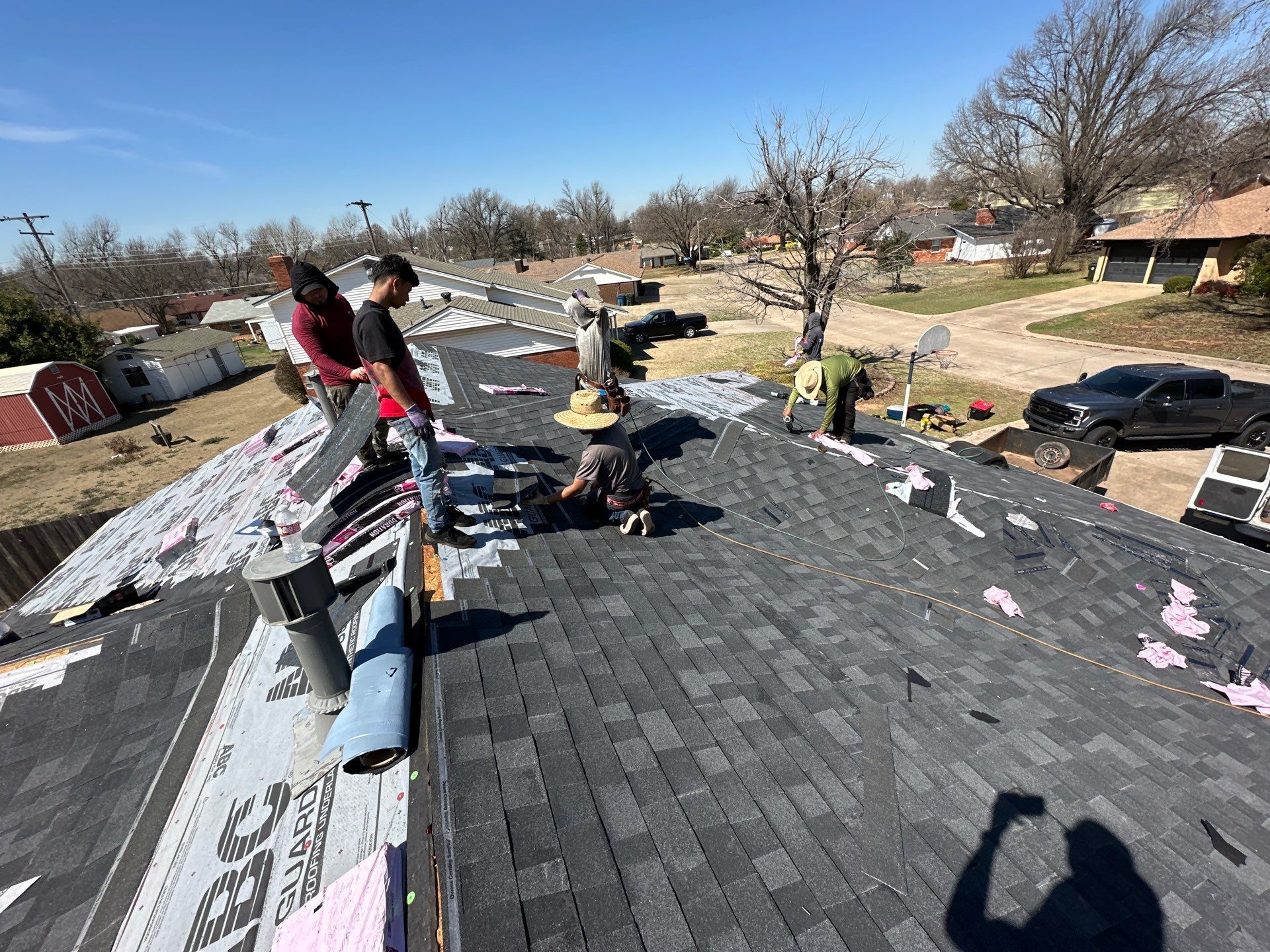 Roofers installing shingles on a house roof on a sunny day. Several workers are visible, with tools, in a residential area.