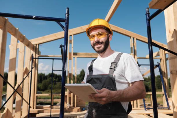 Construction worker in overalls and hard hat holding a tablet, smiling, wooden house frame in background.