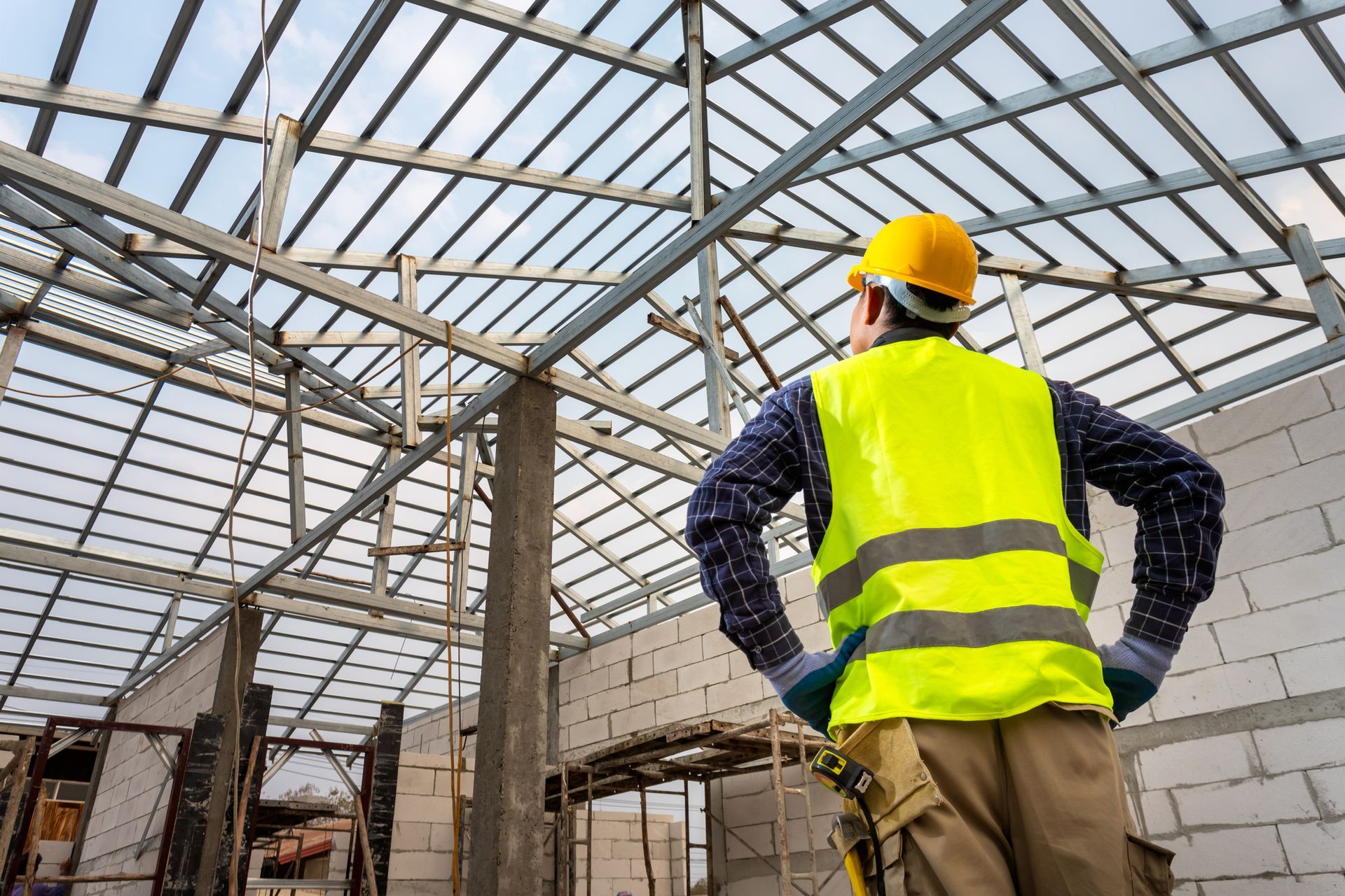 Construction worker in safety vest and hard hat surveying a roof frame under a blue sky.