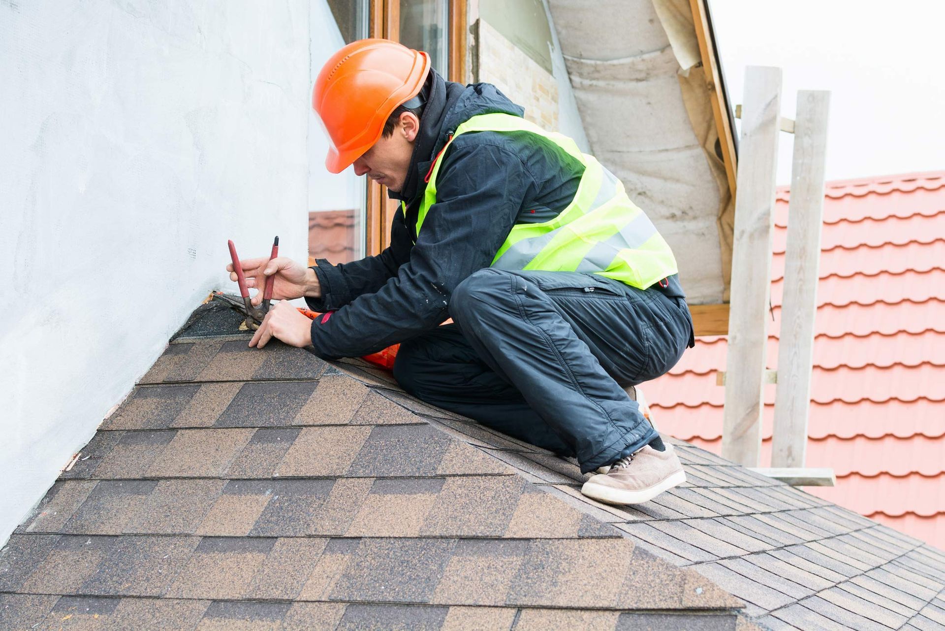 A roofing service contractor wearing protective gear is dismantling old roof shingles.