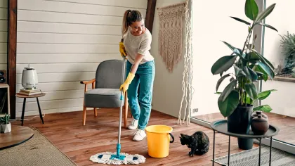Woman mopping a wooden floor wearing yellow gloves. A yellow bucket and black cat are nearby. The room is sunny with plants.