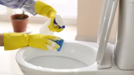 Person wearing yellow gloves cleaning a toilet bowl with a sponge and spray bottle in a bathroom.