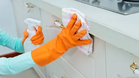 Person wearing orange gloves sprays and wipes a white kitchen cabinet with a cloth.