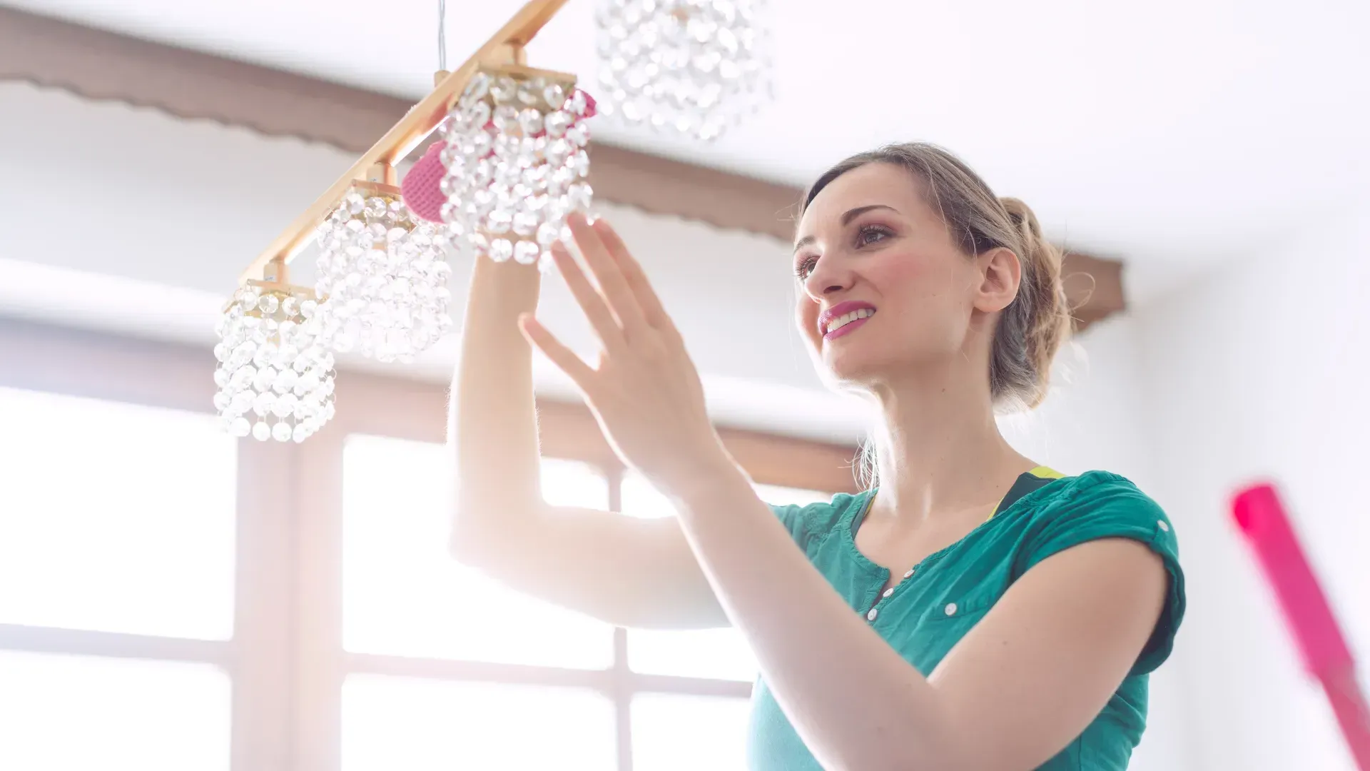 Woman in green shirt smiles while cleaning a crystal chandelier in a bright room with a window and wooden beams.