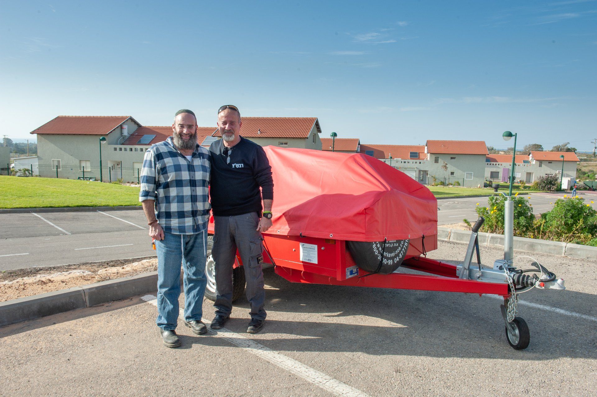 Two men are standing next to a red trailer in a parking lot.