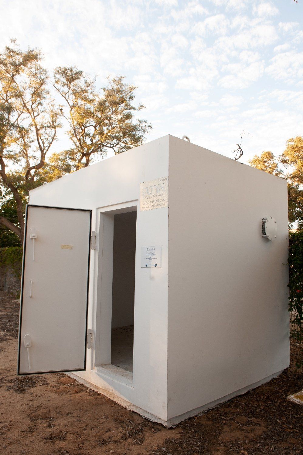 A small white Shelter with a door open and trees in the background.