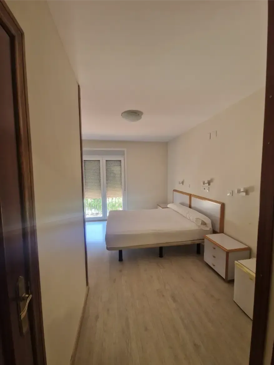 Dormitorio minimalista con cama, mesitas de noche y puertas que dan al balcón, dejando entrar la luz natural.