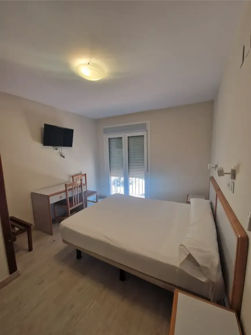 Habitación pequeña con cama, televisor montado en la pared, escritorio y ventana con contraventanas.