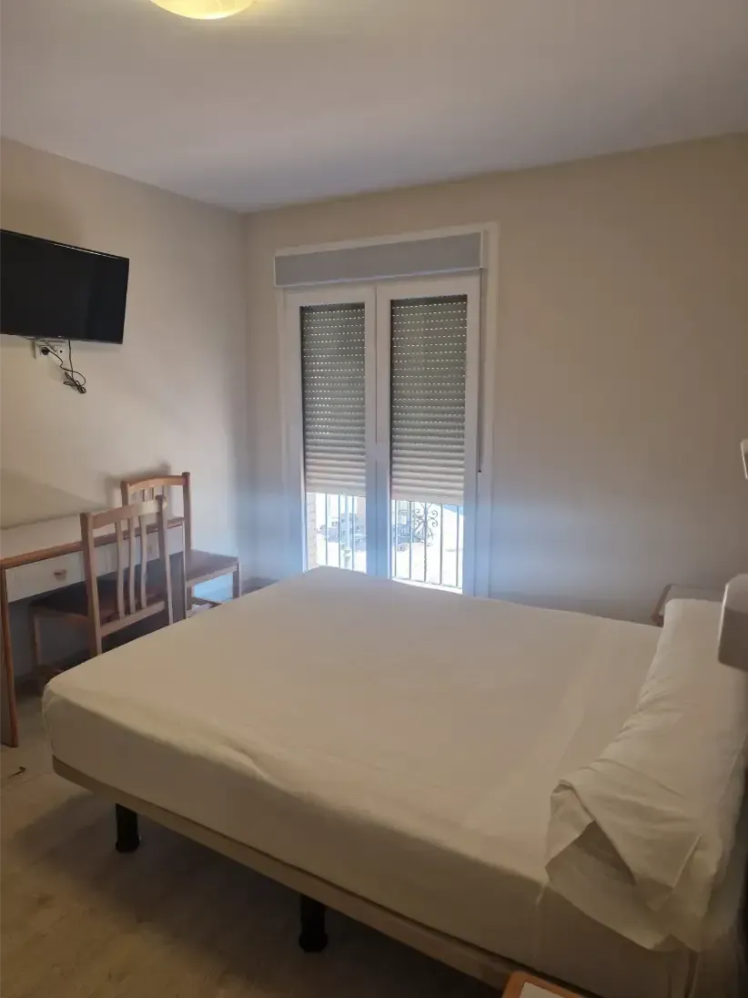 Habitación vacía con cama sin hacer, escritorio, televisor montado en la pared y puertas de balcón con persianas abiertas.