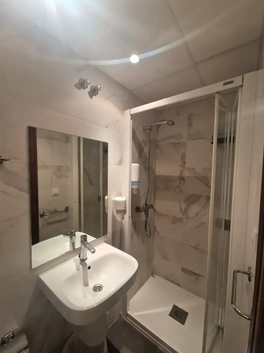 Baño compacto de color blanco con lavabo, espejo, cabina de ducha y puerta de cristal.